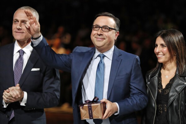 Brooklyn Nets broadcaster Ian Eagle, center, waves to the crowd as Nets' CEO Brett Yormark, left, applauds during a ceremony honoring Eagle's 25-year broadcast career at an NBA basketball game between the New York Knicks and the San Antonio Spurs, Feb. 21, 2019, in New York. Eagle's wife Amy is at right. (AP Photo/Kathy Willens, FIle)
