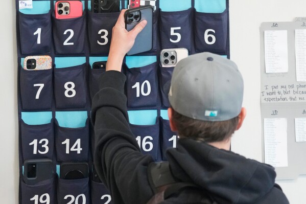 A ninth grader places his cellphone into a phone holder as he enters class at Delta High School, Friday, Feb. 23, 2024, in Delta, Utah. (AP Photo/Rick Bowmer, file)