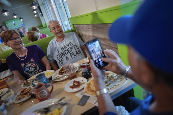 Joe Dubman takes a picture of friends Chris Stanley, wearing an "I have concepts of a plan" T-shirt poking fun at the recent debate performance of Republican presidential nominee former President Donald Trump, and Dee Melvin, wearing a shirt showing support for Democratic presidential nominee Vice President Kamala Harris, as a group of Democratic friends participate in their regular brunch meet-up, in The Villages, Fla., Sunday, Sept. 15, 2024. (AP Photo/Rebecca Blackwell)